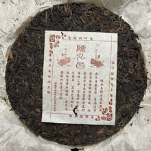 Load image into Gallery viewer, Compressed round puerh tea cake with Chinese label on parchment paper, loose tea leaves visible