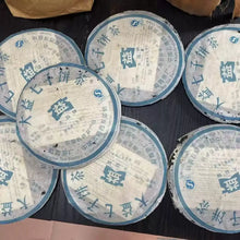 Load image into Gallery viewer, Seven compressed puerh tea cakes in blue and white wrappers on a dark table