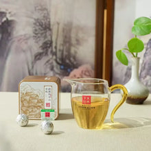 Load image into Gallery viewer, Glass pitcher of Chinese tea, metal tea tin, and wrapped tea balls on table with plant
