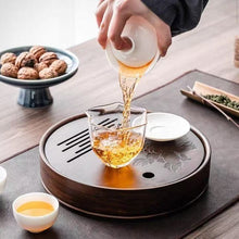 Load image into Gallery viewer, Tea being poured from a teapot into a glass cup on a tea tray with a wooden surface.  Product Name: Round Tea Tray

Material: Bamboo + Laminate Board Base

Size:

Small : Diameter 25 * Height 5 cm

Medium: Diameter 29 * Height 5 cm

Large : Diameter 38 * Height 5 cm



This Round Tea Tray is an exquisite piece of craftsmanship, designed for the ritual of Gongfu tea, a traditional Chinese method of tea preparation that focuses on the aesthetic presentation and sharing. The tray is constructed from high-quali