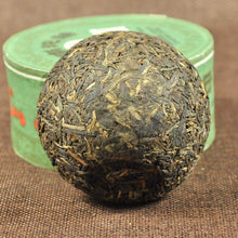 Carica l'immagine nel visualizzatore di Gallery, Compressed Chinese Puerh tea ball in front of green tea tin on brown fabric