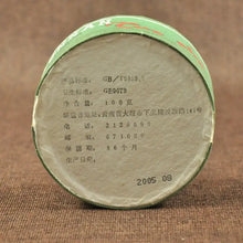 Carica l'immagine nel visualizzatore di Gallery, Round puerh tea cake package with Chinese text and 2005 production date on brown fabric background