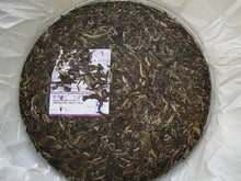 Load image into Gallery viewer, Compressed pu-erh tea cake with visible tea leaves and a label, wrapped in paper.