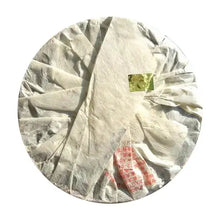 Carica l'immagine nel visualizzatore di Gallery, Round compressed puerh tea cake wrapped in white paper with red Chinese text, Chinese tea.