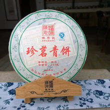 Load image into Gallery viewer, Chinese raw pu-erh tea cake, wrapped in paper with green and red Chinese characters, on a wooden stand