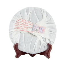 Load image into Gallery viewer, Chinese puerh tea cake wrapped in white paper on a wooden stand with red Chinese label