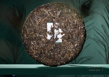 Load image into Gallery viewer, Compressed puerh tea cake displayed upright against a green background with peacock feather pattern.