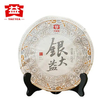 Load image into Gallery viewer, Round Chinese puerh tea cake wrapped in decorative paper on a black display stand