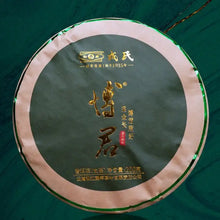 Load image into Gallery viewer, Compressed round puerh tea cake in green and tan wrapper with Chinese characters on table.