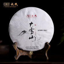 Load image into Gallery viewer, Chinese pu-erh tea cake wrapped in white paper with mountain artwork, displayed on a wooden stand