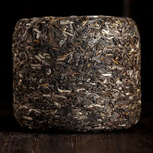 Load image into Gallery viewer, Close-up of a compressed sheng puerh tea brick with visible tea leaves on a dark wooden surface